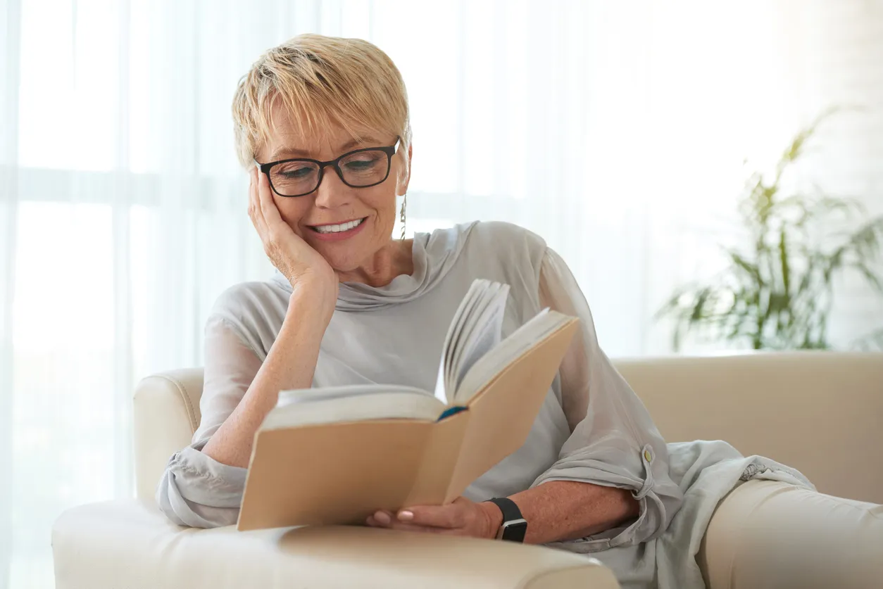 Ältere Dame mit Brille liest ein Buch auf dem Sofa