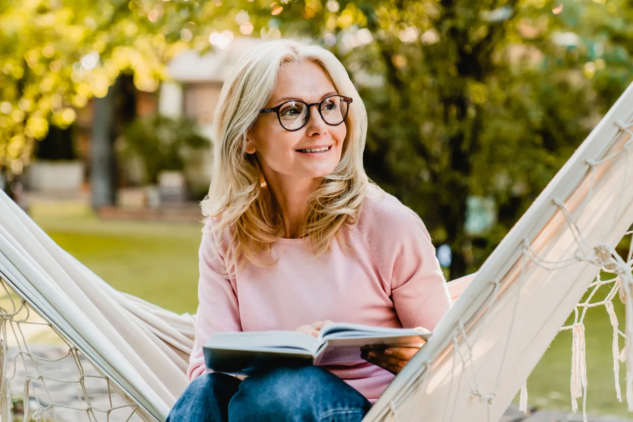 Seniorin mit Brille und Buch auf Hängematte im sommerlichen Garten